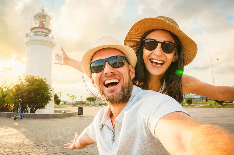 Young excited caucasian couple in love smiling taking selfie pov at Batumi Boulevard with iconic lighthouse and Ferris wheel during golden hour. Happy couple Holiday taking photo by popular landmarks