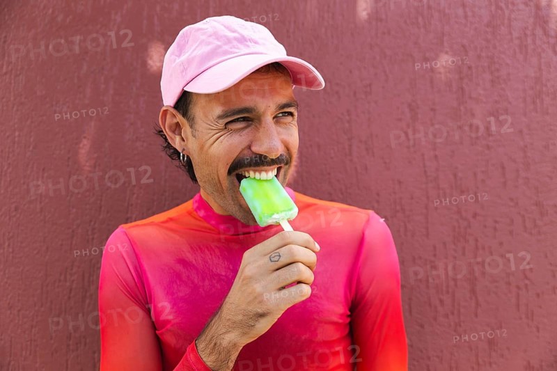 A young Spanish LGTBIQ+ man with a mustache showcases lively urban fashion as he enjoys a popsicle in Quito, Ecuador