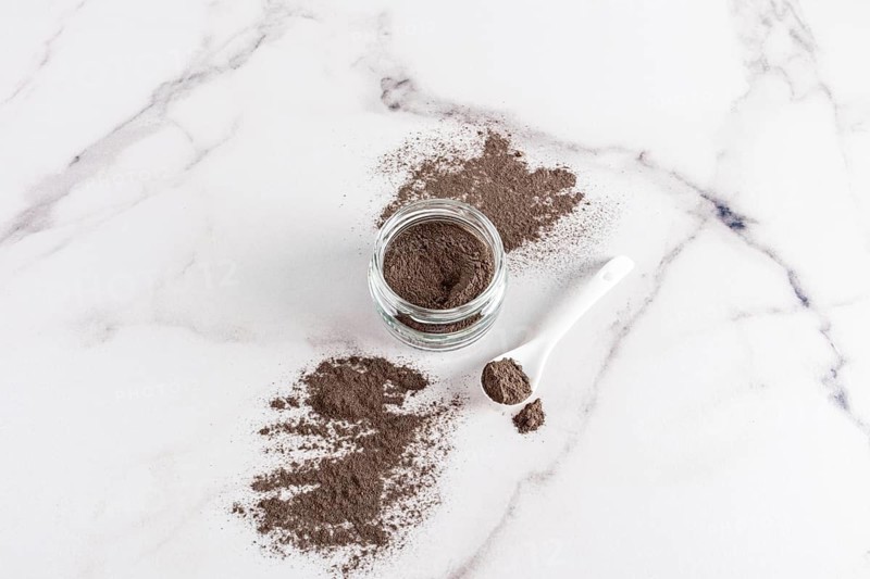Top view of open glass jar filled with cosmetic clay powder, accompanied by a white spoon on a sleek marble surface, ideal for skincare routines