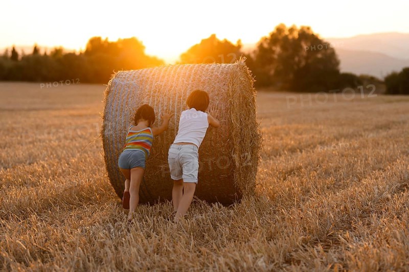 Back view of two unrecognizable children, a boy and a girl, play by pushing a large hay bale in a field during sunset, creating a warm, golden scene