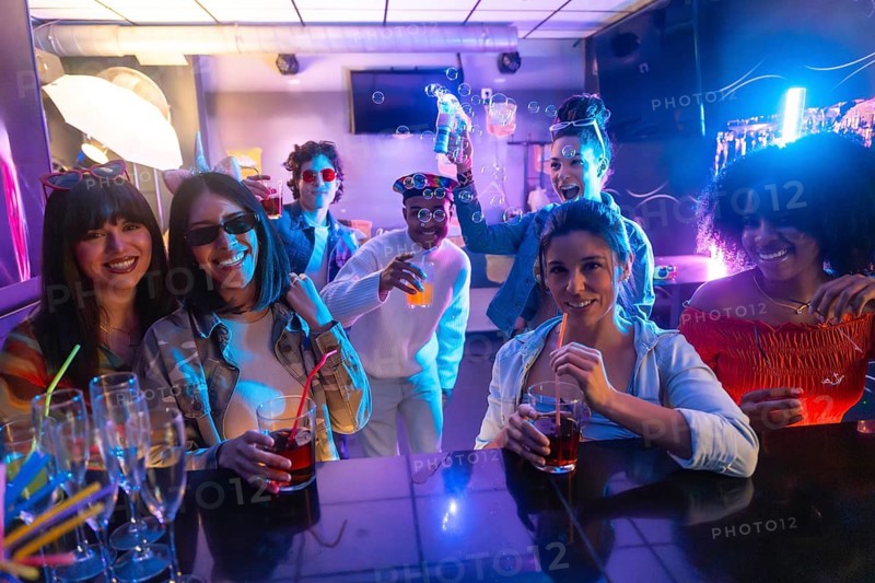 Group of diverse young adults laughing, smiling, and enjoying drinks and bubbles at a vibrant nightclub, celebrating a special occasion with illuminated lights