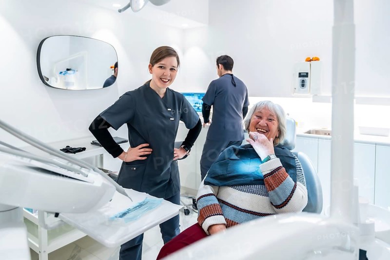 Happy elderly patient sitting in dentist chair while doctor and nurse smiling in modern bright dental clinic