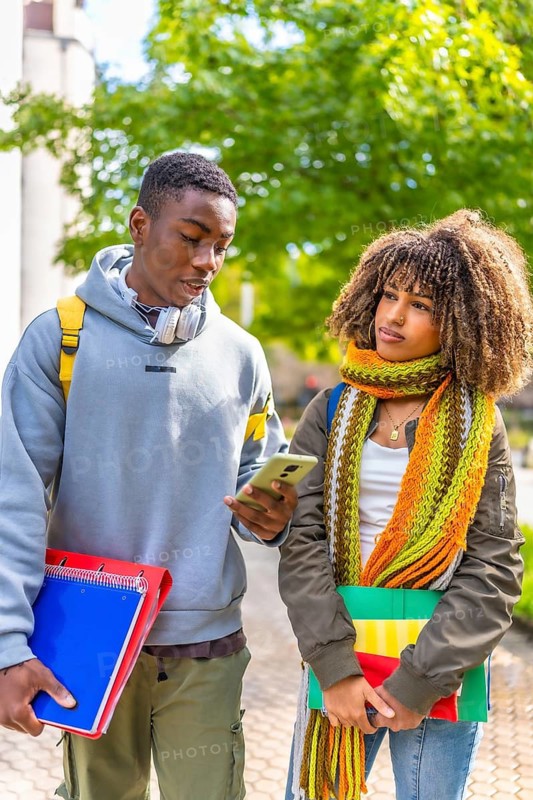 Vertical photo of two multi-ethnic latin and african students using mobile phone while walking along the university campus