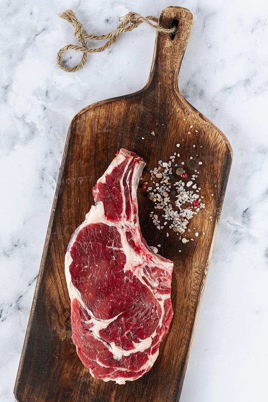 Raw rib eye steak, cowboy steak, wooden chopping board, marble table, top view, no people