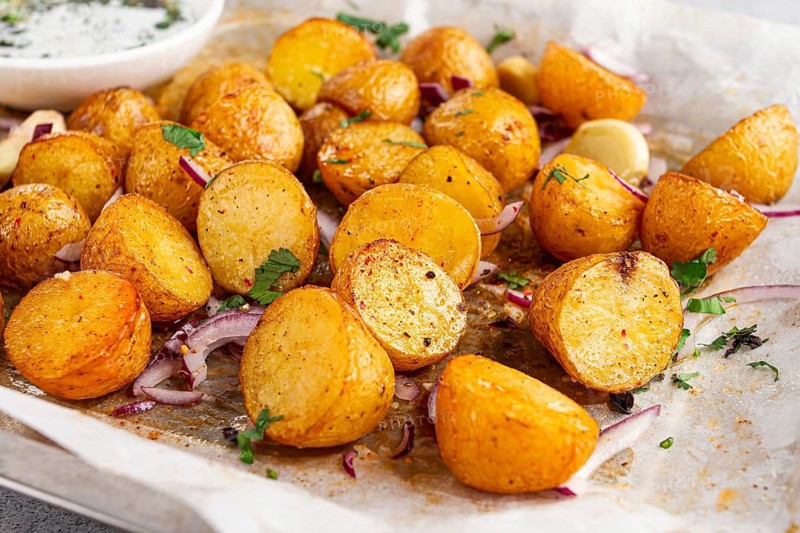 Roasted potatoes sit on a baking sheet next to chopped herbs and red onions. They are golden brown and ready to be served. Ingredients add color and flavor to the dish