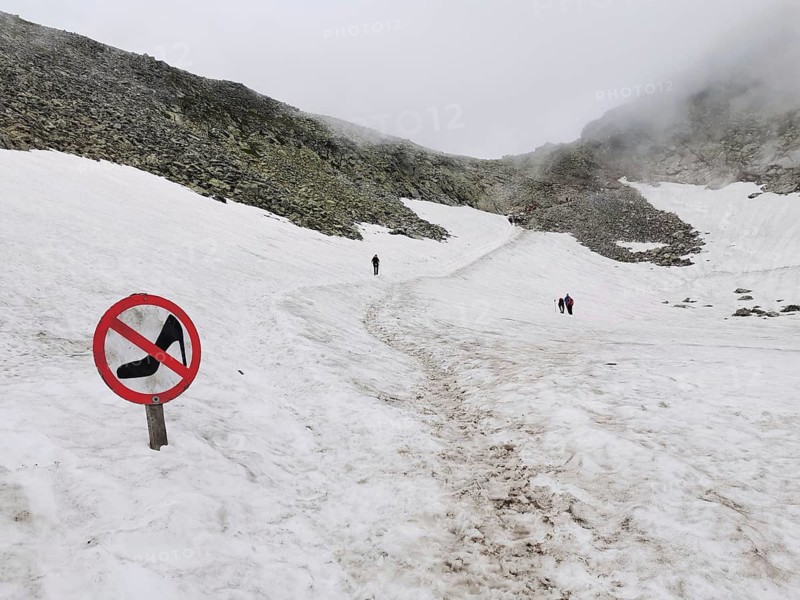 People walk across a snow-covered mountain field with warning sign, no high heels, high heels, funny, in foggy conditions, hiking, Rysy, High Tatras, Slovakia