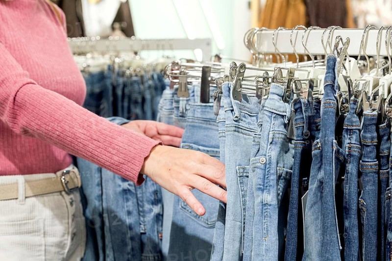 A woman in a pink sweater selects blue jeans from a clothing rack. The scene captures the retail shopping experience with a focus on style and fashion choices