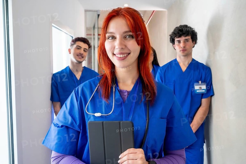 Young red haired medical student with stethoscope smiling confidently in front of a diverse healthcare team in blue scrubs, standing in a clinic hallway, ready to care