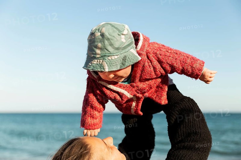 A mother joyfully lifts her child at the beach in Samalus, Barcelona. The toddler wears a cozy red sweater and green hat, capturing a moment of love and freedom