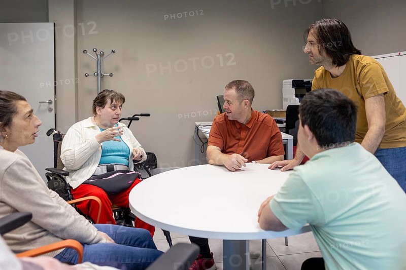 People gather around a table in a supportive day center. They engage in discussion, highlighting community and empowerment for individuals with mental disabilities
