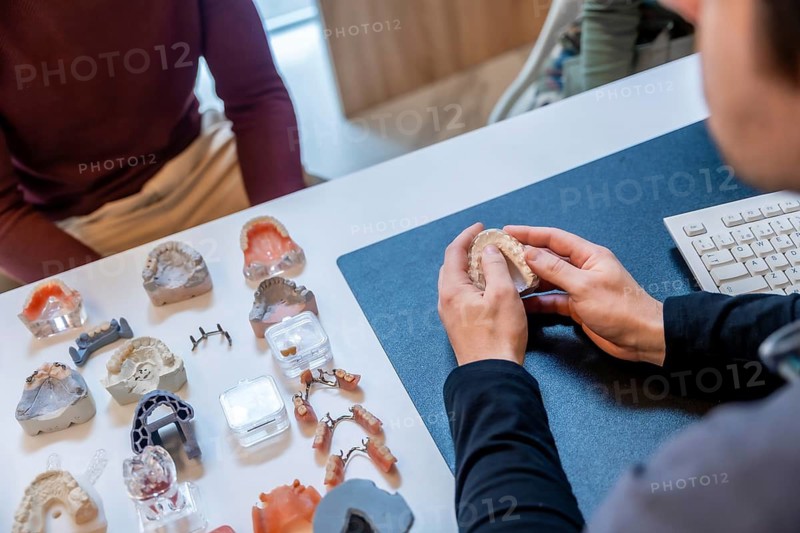 Dentist explaining to patient treatment with dental prosthesis, surrounded by various dental models in clinic