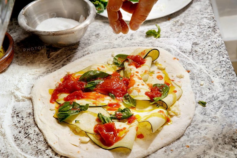 Cropped unrecognizable person hands adds toppings to a fresh pizza dough with zucchini slices, basil, cheese, and tomato sauce on a granite counter