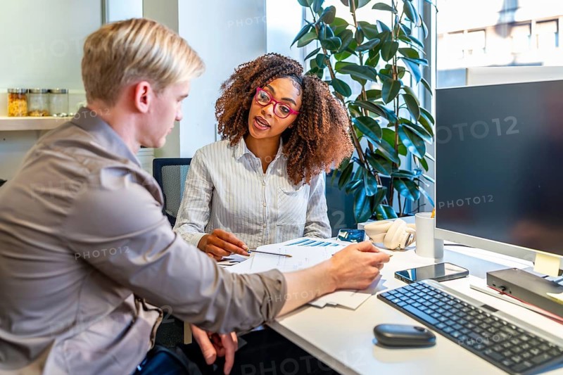 Two coworkers, a man and a woman, are discussing charts and data in a modern coworking office, collaborating on a project
