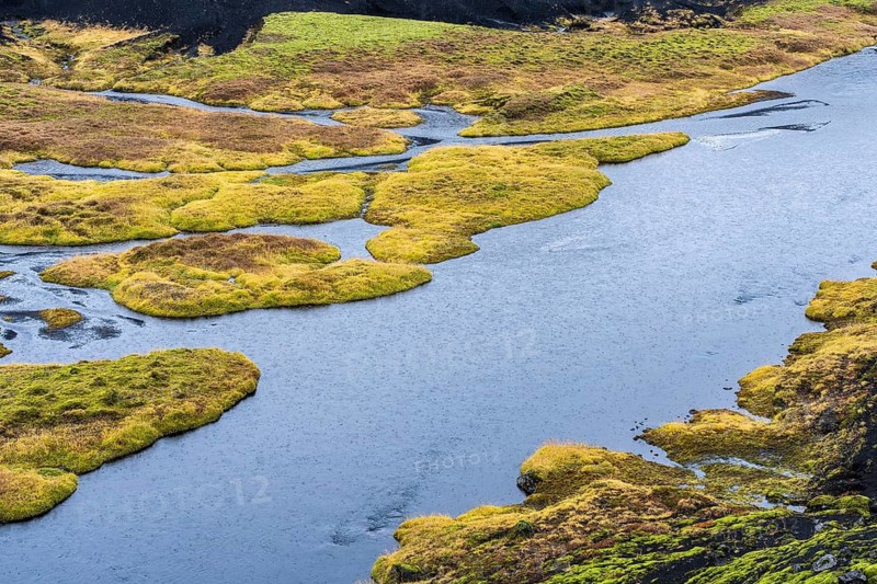 An aerial view of an Icelandic riverside captures dynamic water flow surrounded by vibrant green moss and unique earth textures, showcasing the natural beauty and contrast of the landscape