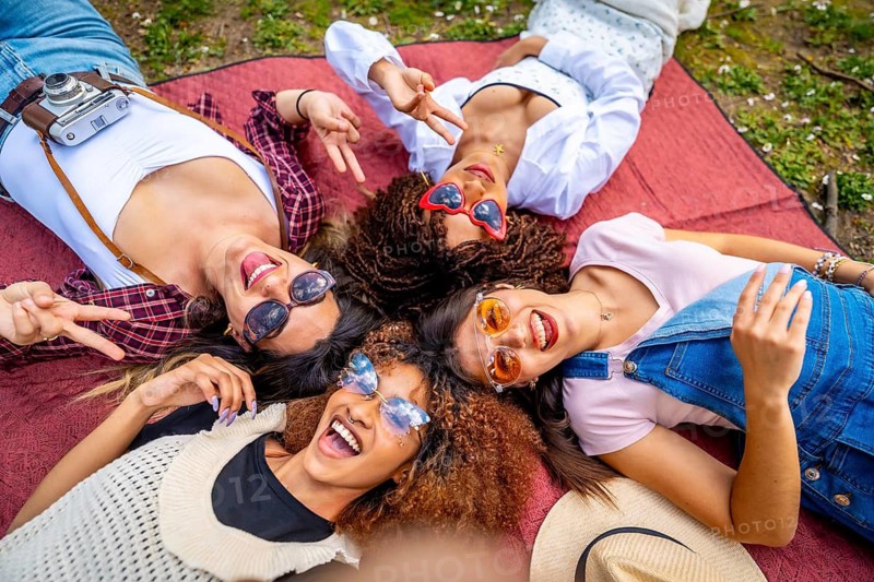 Four multi ethnic female friends are lying on a blanket in a park, making a selfie and gesturing peace signs