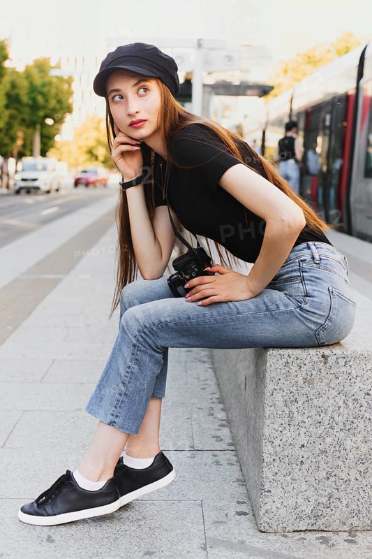 A young woman with long hair and casual attire sits on a city bench, holding a camera. She is surrounded by urban scenery, featuring a tram and passing cars