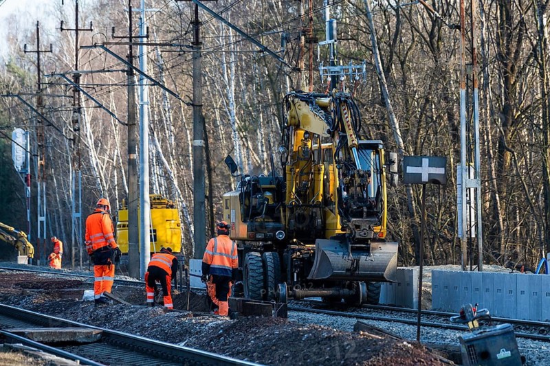 Railway construction crew in high-visibility gear working on tracks alongside a yellow road-rail excavator during infrastructure maintenance near a forest in Tymowa, Poland