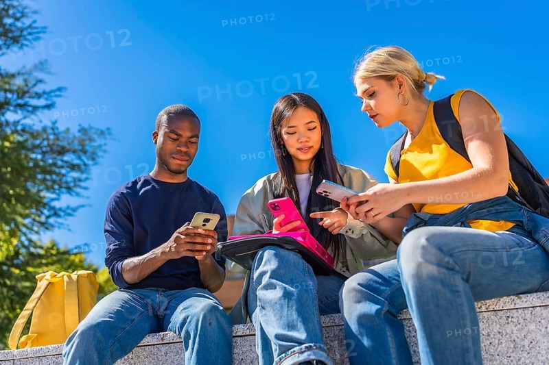 Low angle view photo of a multi-ethnic group of students using mobile phone sitting on campus bench