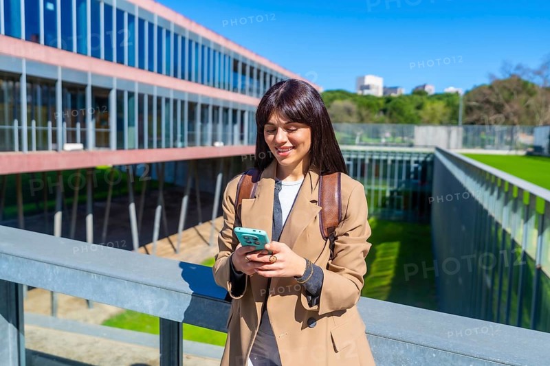 Smiling young woman standing outdoors on a university campus, checking her smartphone while wearing a backpack and casual attire, embodying student life and connected education