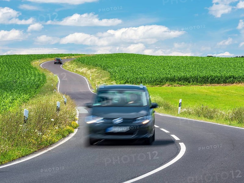 Car driving on winding country road through green fields under blue sky with white clouds in spring, Thuringia, Germany