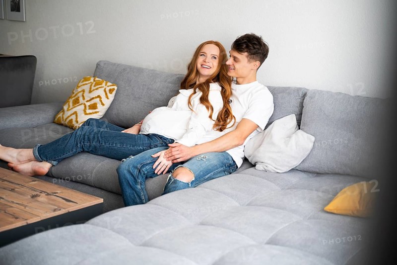 Young couple sitting on a cozy sofa at home, expecting a baby. They share a joyful and loving moment, surrounded by cushions, embodying warmth and togetherness