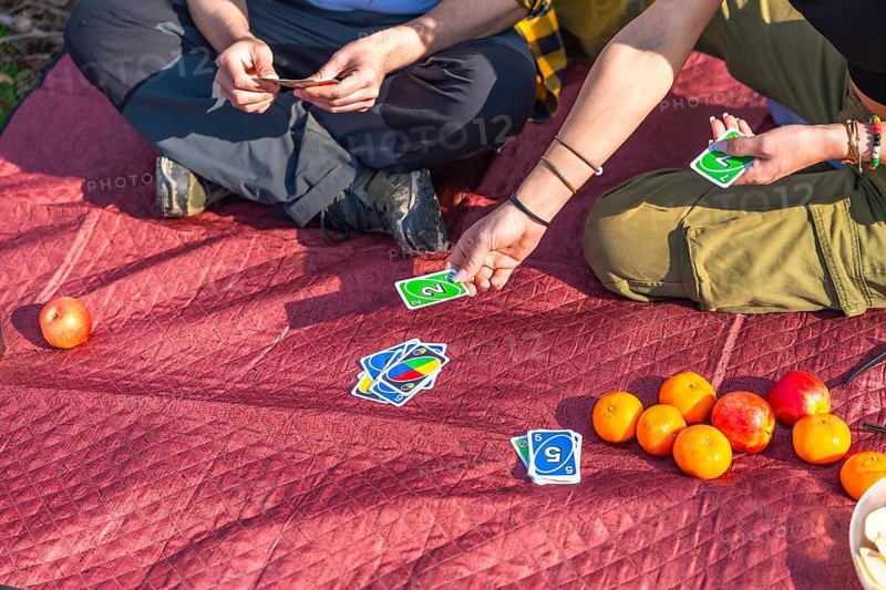 Hikers enjoying a lively card game while relaxing on a red quilted blanket, surrounded by fresh apples and oranges during a sunny picnic