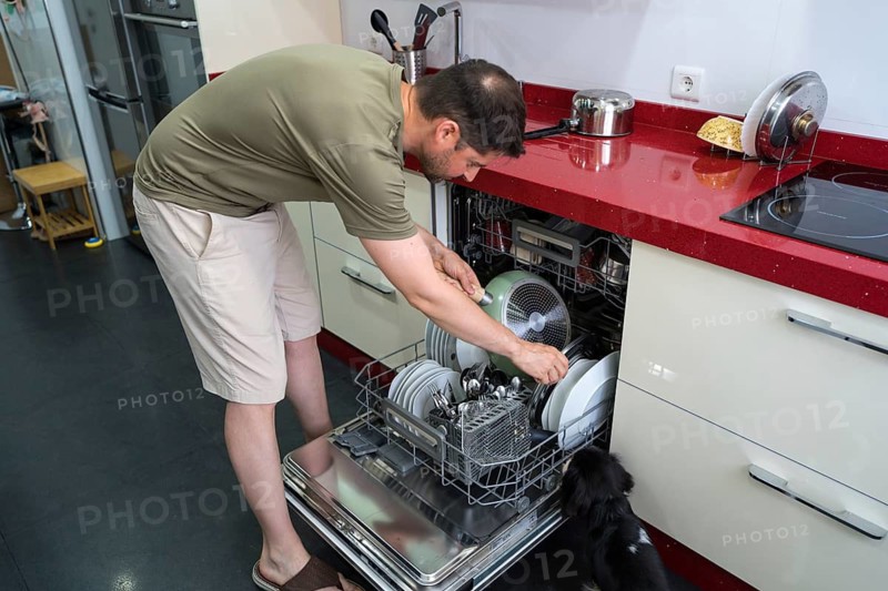 A man in casual attire loads dishes into a dishwasher in a modern kitchen. The countertop features red accents and kitchen utensils, while a curious dog observes the chore