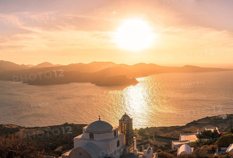 Sun sets behind Greek island, casting golden hues over the Aegean Sea. A white domed church is silhouetted against the vibrant sky, creating a serene, picturesque scene in Plaka, Milos island