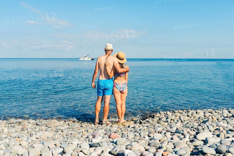 Caucasian Man and woman in summer hats embracing while standing on sunny pebble beach facing sea horizon, concept of romance, wellness, well being relaxation, travel seaside holiday. Analog film look