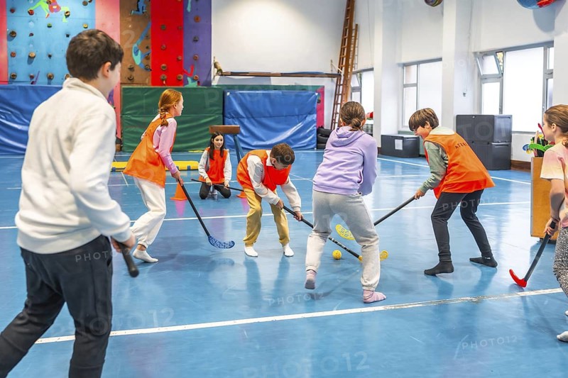Group of children wearing bright orange vests, actively playing floorball together in a lively school gym, enjoying teamwork and fun