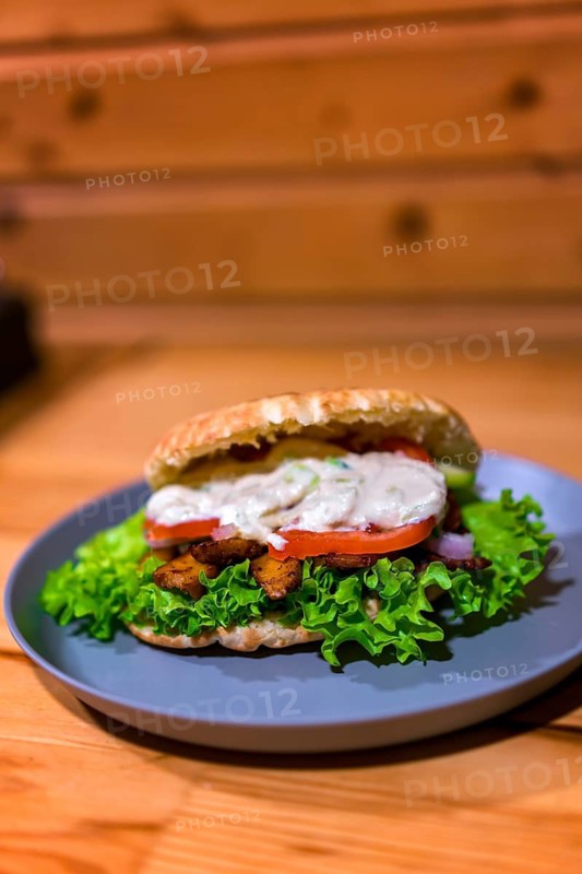 Close-up of a Greek-style sandwich with fresh lettuce, tomatoes, seasoned meat, and creamy sauce on a gray plate. Served on a wooden table, perfect for fast-food lovers
