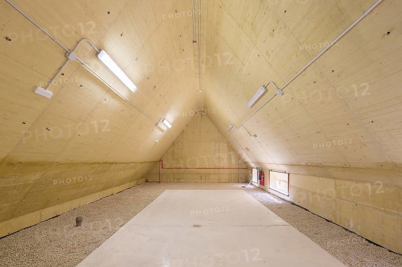 Large industrial attic of hospital with an unfinished interior showcasing exposed wooden beams. Concrete floor and gravel surface. Adequate lighting for a raw architectural look
