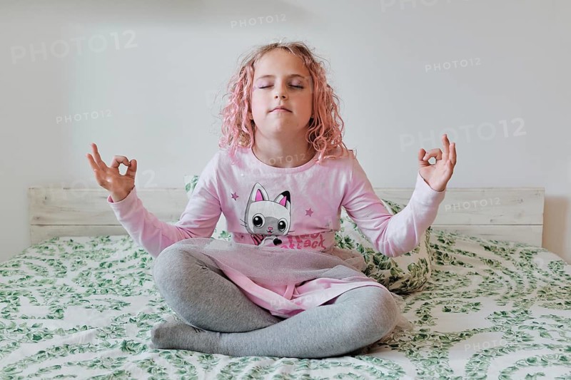 A young girl with pink hair sits cross legged on a bed, eyes closed, practicing meditation. She wears a pink cat themed outfit, embodying serenity in a cozy home setting
