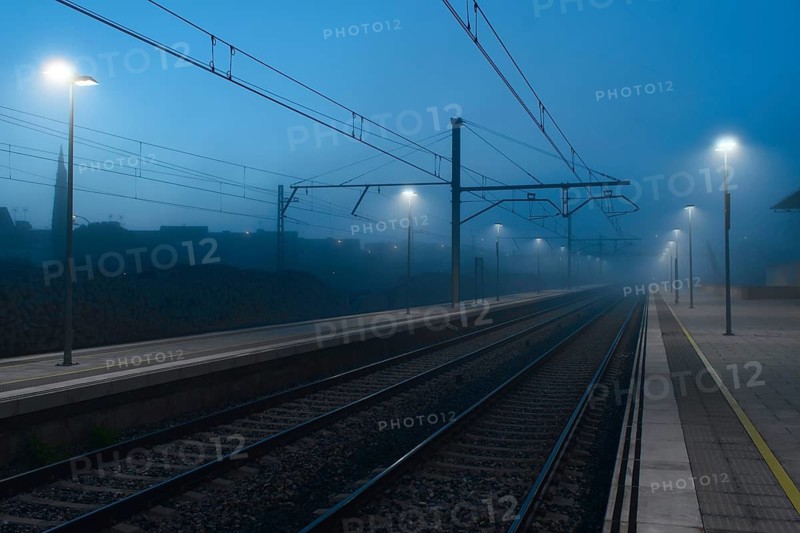 A serene railway station engulfed in fog under the dim glow of street lights, with empty tracks extending into the misty distance