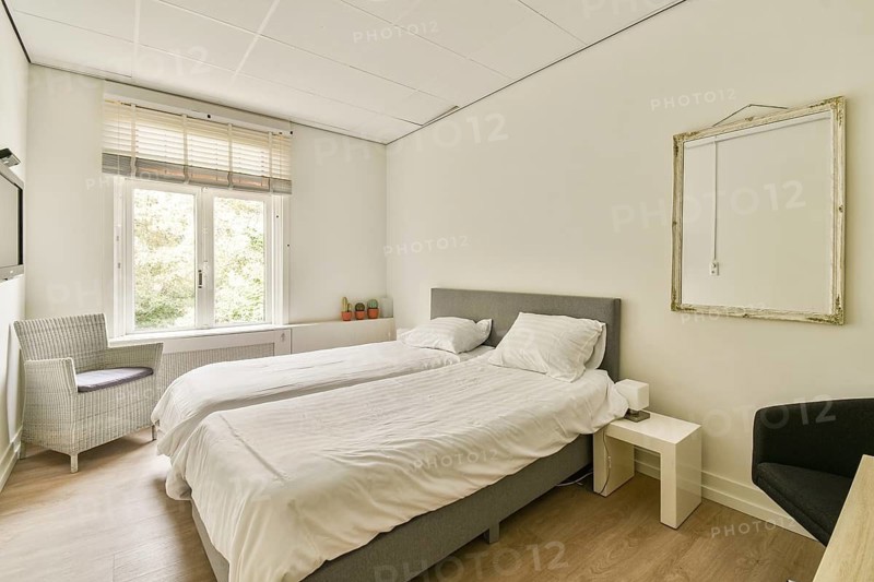 A serene, modern bedroom featuring twin beds, a stylish armchair, a sleek mirror, and natural light streaming through the windows, creating an inviting atmosphere