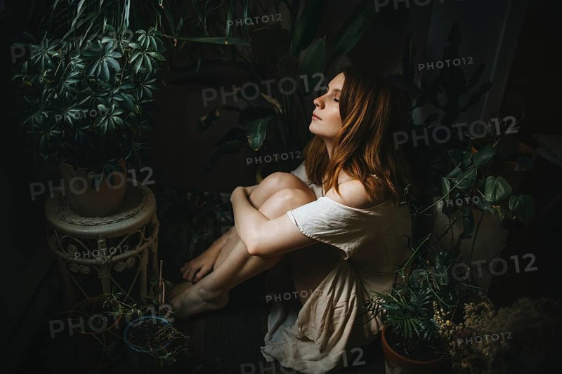 A serene scene of a woman sitting among lush indoor plants, bathed in soft light. The ambiance is calm and reflective, embodying peace and connection with nature