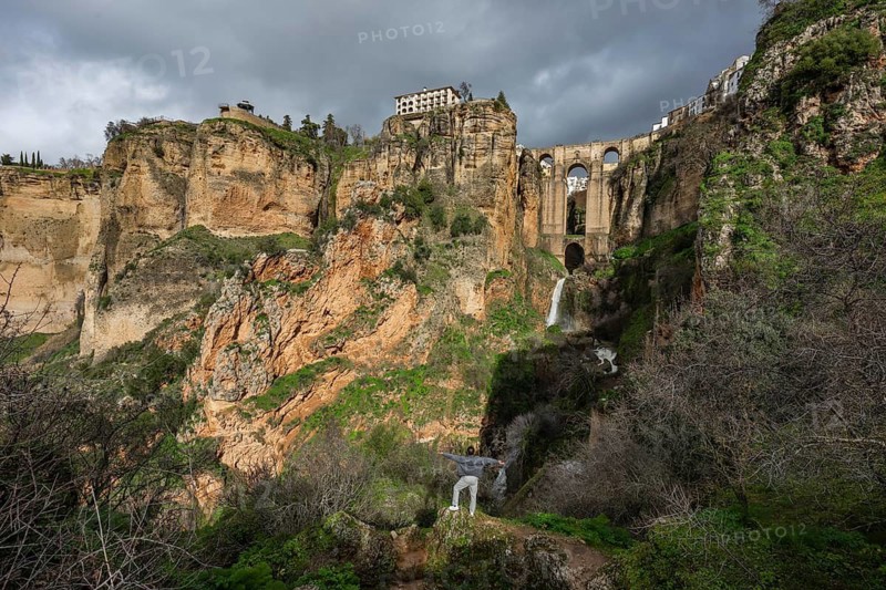 A traveler stands on a rock overlooking a waterfall and the massive arches of the Puente Nuevo bridge from the bottom of the gorge in Ronda, Spain