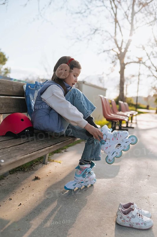 Side view of a young girl sits on a wooden bench in a sunlit park, putting on her roller skates, with a red helmet nearby