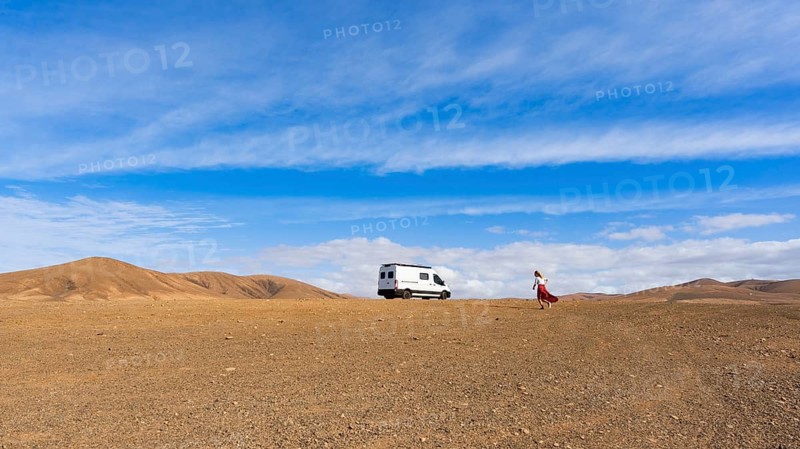 A white van is parked in the vast, arid desert of Fuerteventura, Canary Islands, Spain. A woman in a red skirt walks towards the horizon under a bright blue sky