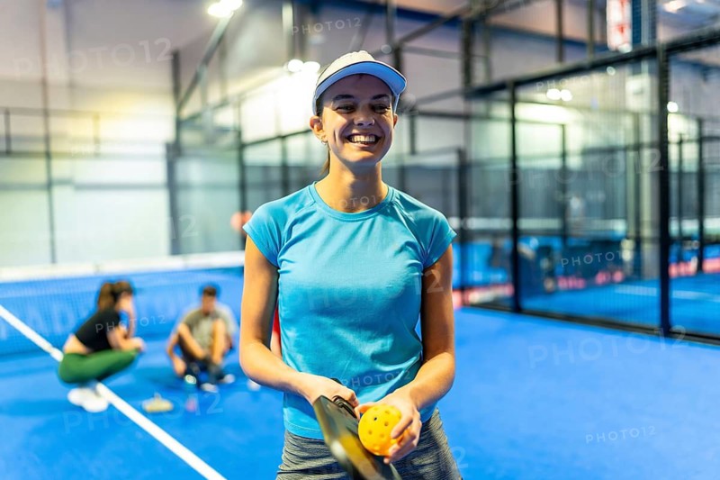 Woman smiling at camera while holding a pickleball paddle and ball on a blue indoor court, teammates relaxing in background enjoying casual, energetic recreational play