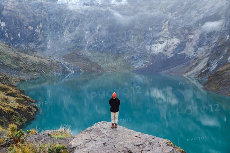 An Ecuadorian woman stands on a rocky ledge overlooking a serene turquoise lake at El Altar Volcano in Ecuador, surrounded by majestic mountainous scenery