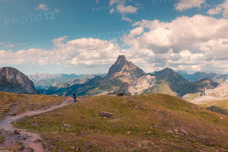 A lone hiker walks along a scenic trail with a stunning mountain peak in the Pyrenees. The expansive sky filled with fluffy clouds creates a serene atmosphere in summer