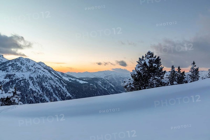 A tranquil mountain scene at sunset featuring pristine snow and dense pine trees against a backdrop of soft sky gradients from orange to blue