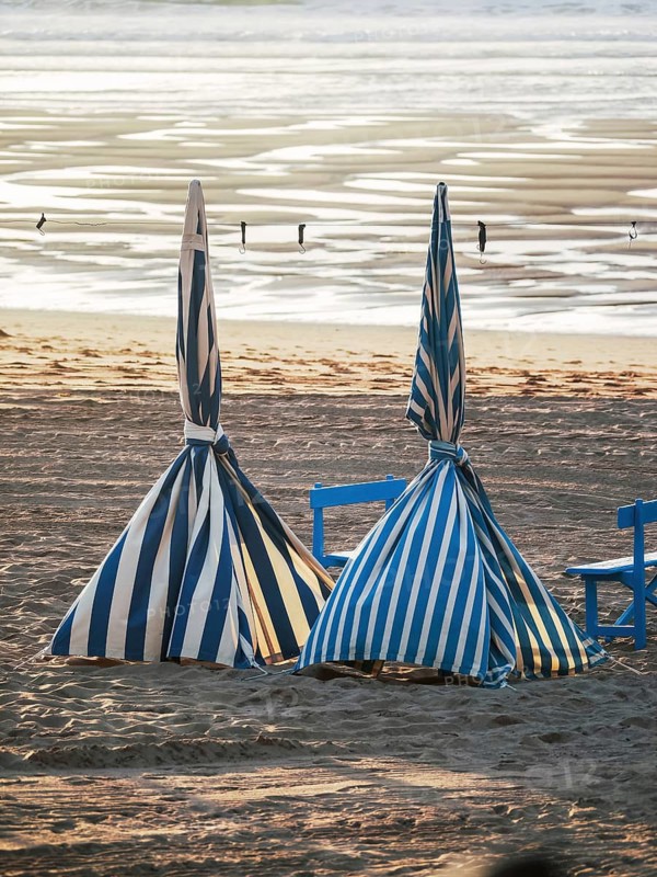 Two striped beach umbrellas are tightly wrapped and unattended on a sandy shore, against a backdrop of reflective water under the sunset