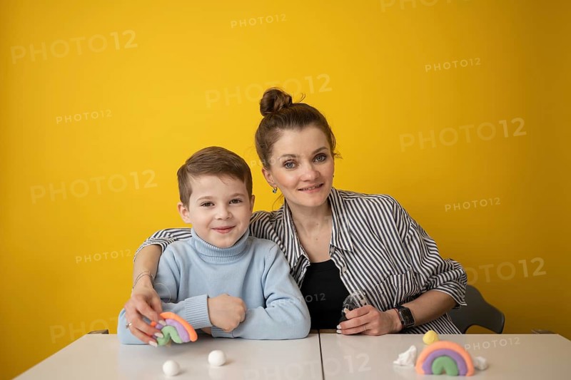 A child and a woman smile in front of a yellow wall, enjoying a creative lesson with colorful modeling clay. They craft rainbows, fostering creativity and bonding through play