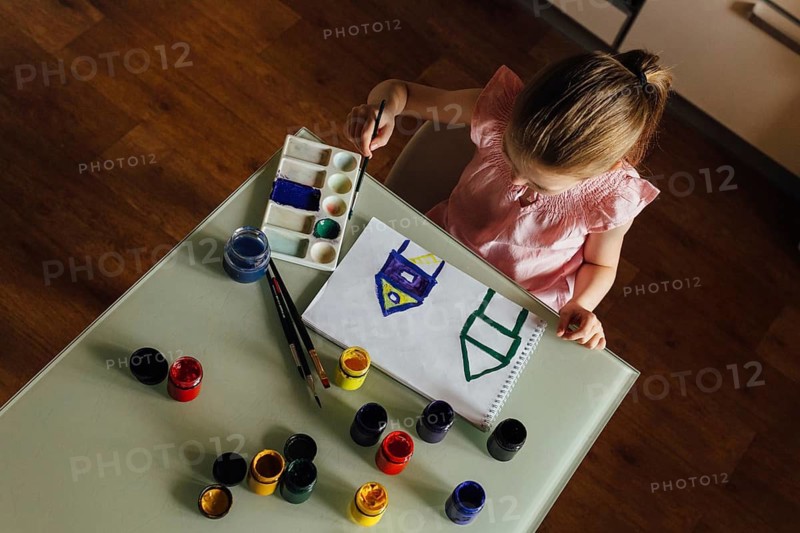 A young girl sits at a table, focused on painting a colorful picture in a sketchbook. Surrounded by paint jars and brushes, she expresses her creativity and imagination