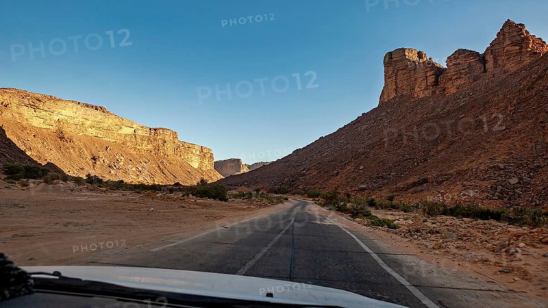 A deserted road leads through a desert landscape with imposing rock formations under a clear blue sky, A drive through the mountainous landscape of the Sahara in Algeria