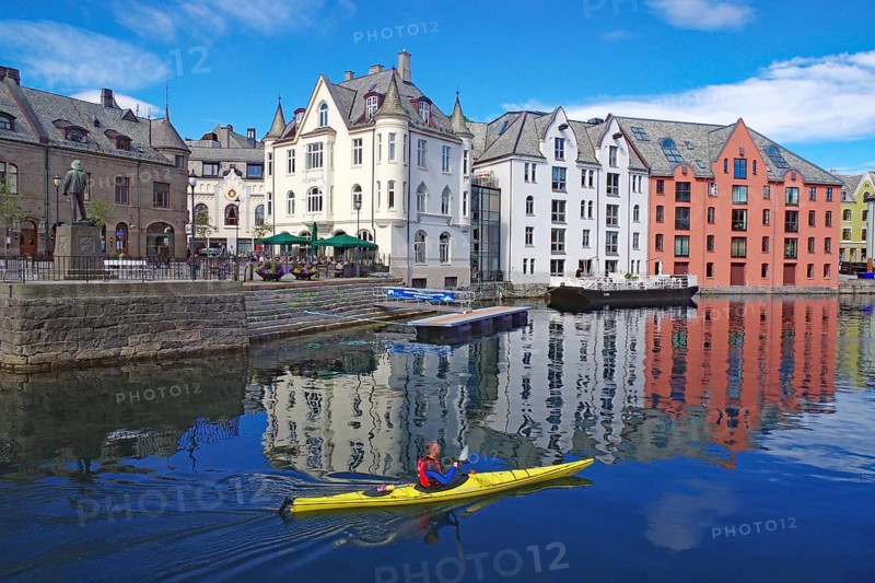 A kayaker paddles past colorful buildings that are reflected in the water in a harbor, Art Nouveau, Alesund, Norway