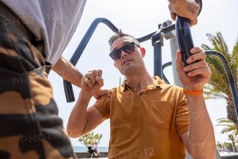 A man assists another in using outdoor exercise equipment on a sunny day. The helper, in a brown shirt and sunglasses, grips the equipment and his companion's hand. The focus is on their interaction and the equipment