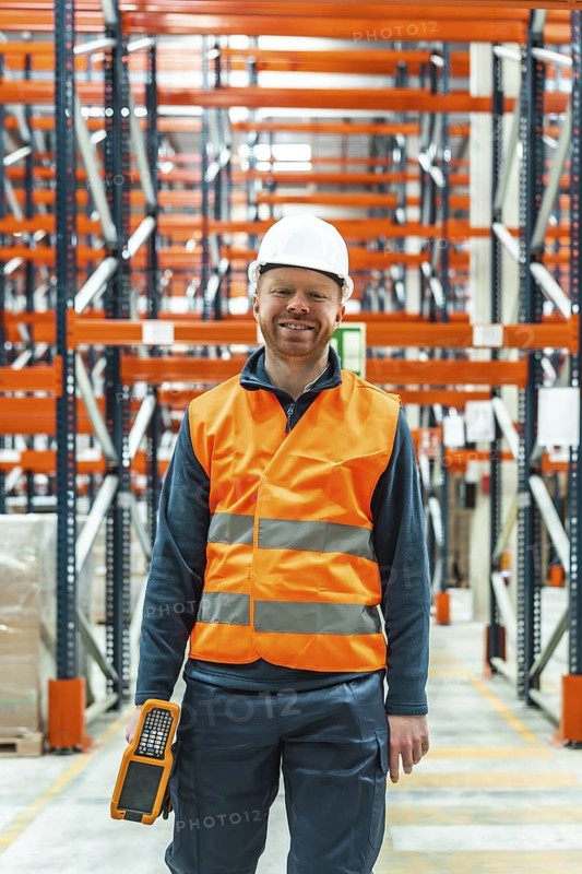 Warehouse worker wearing safety vest and helmet smiling and holding scanner while walking in logistics center
